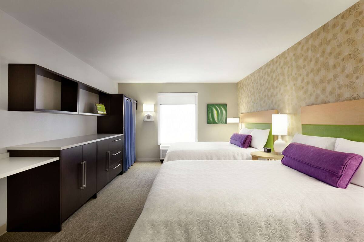A hotel room with two beds, a desk with drawers, a window with curtains, and a wall-mounted lamp.