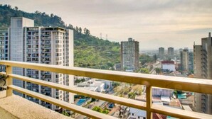 Um edifício alto com vários andares, uma varanda com guarda-corpo e vista para um horizonte urbano com outros edifícios e áreas verdes ao fundo.