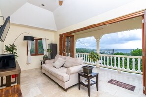 A living room with a beige sofa, a TV on a wooden table, and a balcony with a white railing and potted plants. - Sandrati Villa (Bequia Island)