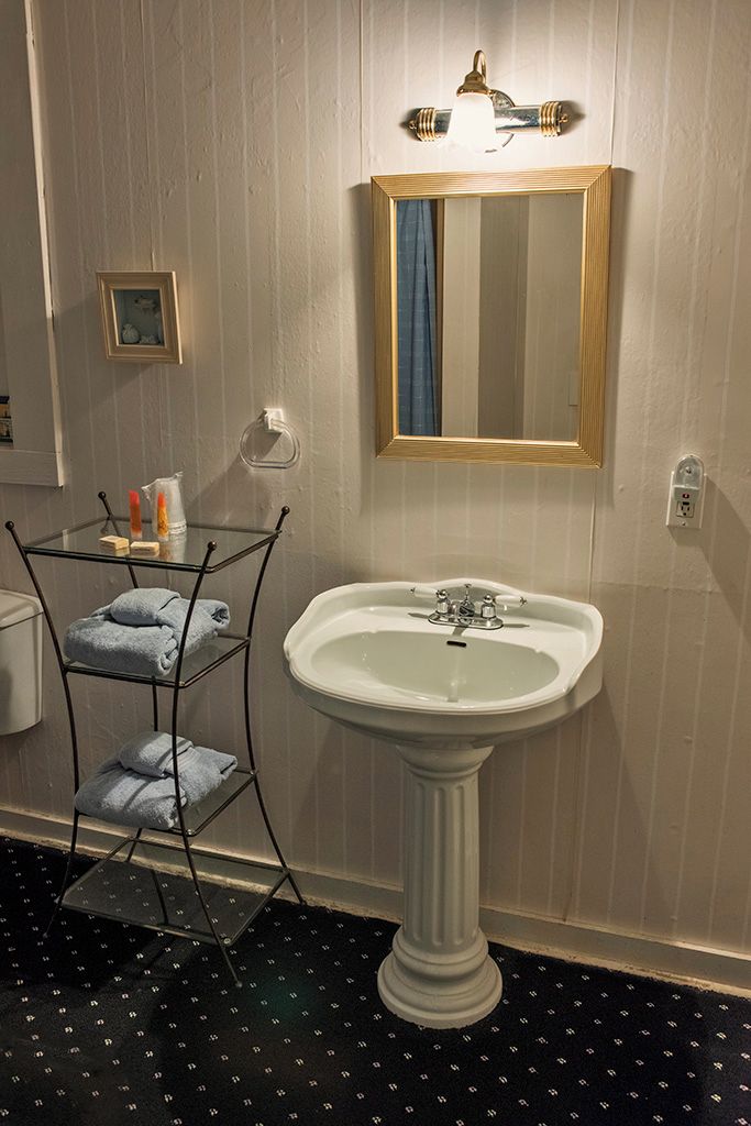 Ett badrum med ett handfat på en piedestal, en handdukshängare med hopvikta handdukar, en inramad spegel och en väggmonterad lampa.