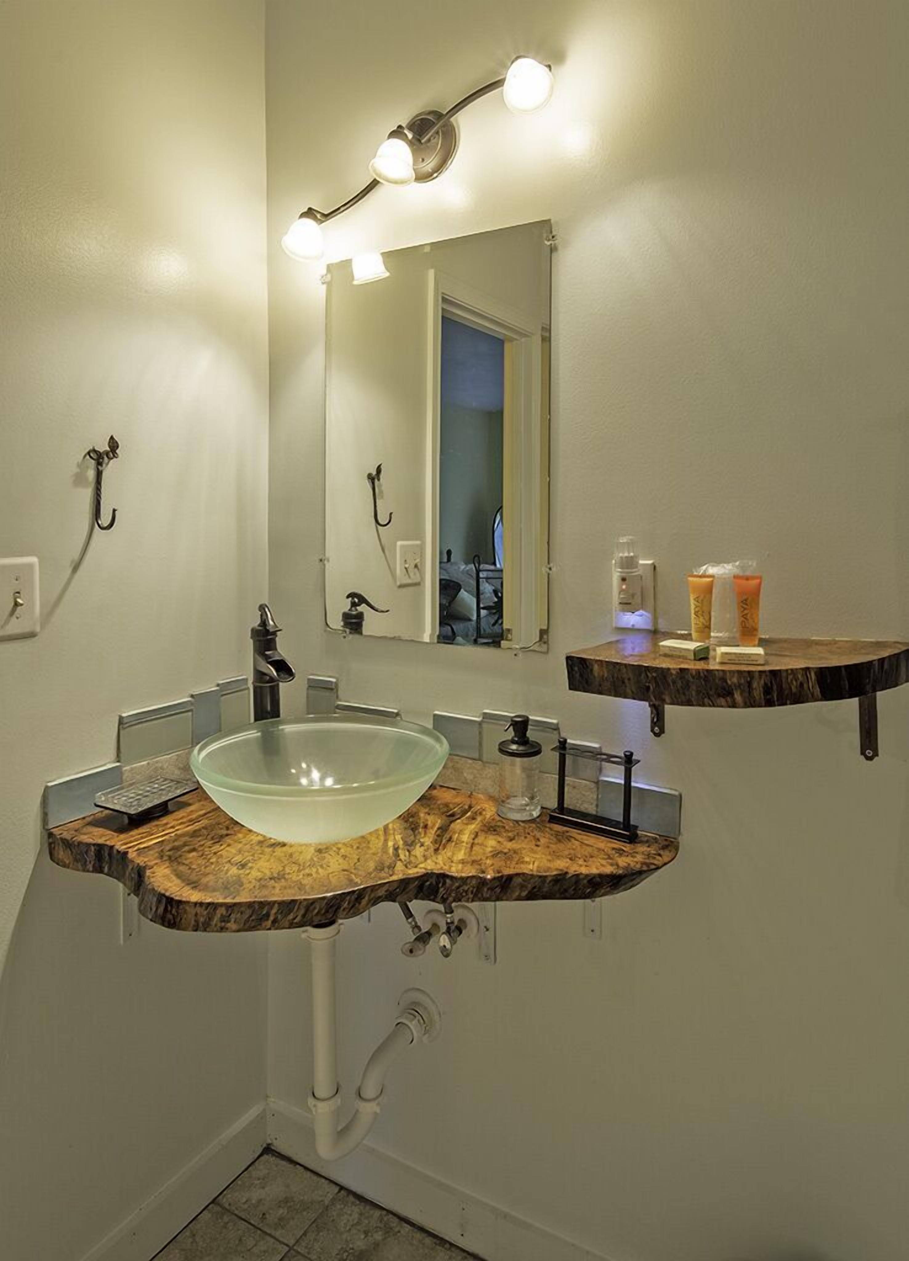 A bathroom with a marble countertop, a glass sink, a mirror with lights, a towel rack, and toiletries on a shelf.