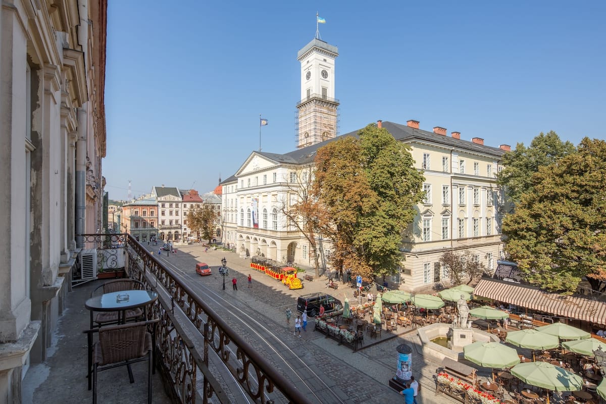 A balcony with a table and chairs, overlooking a street with buildings, a clock tower, and a tram.