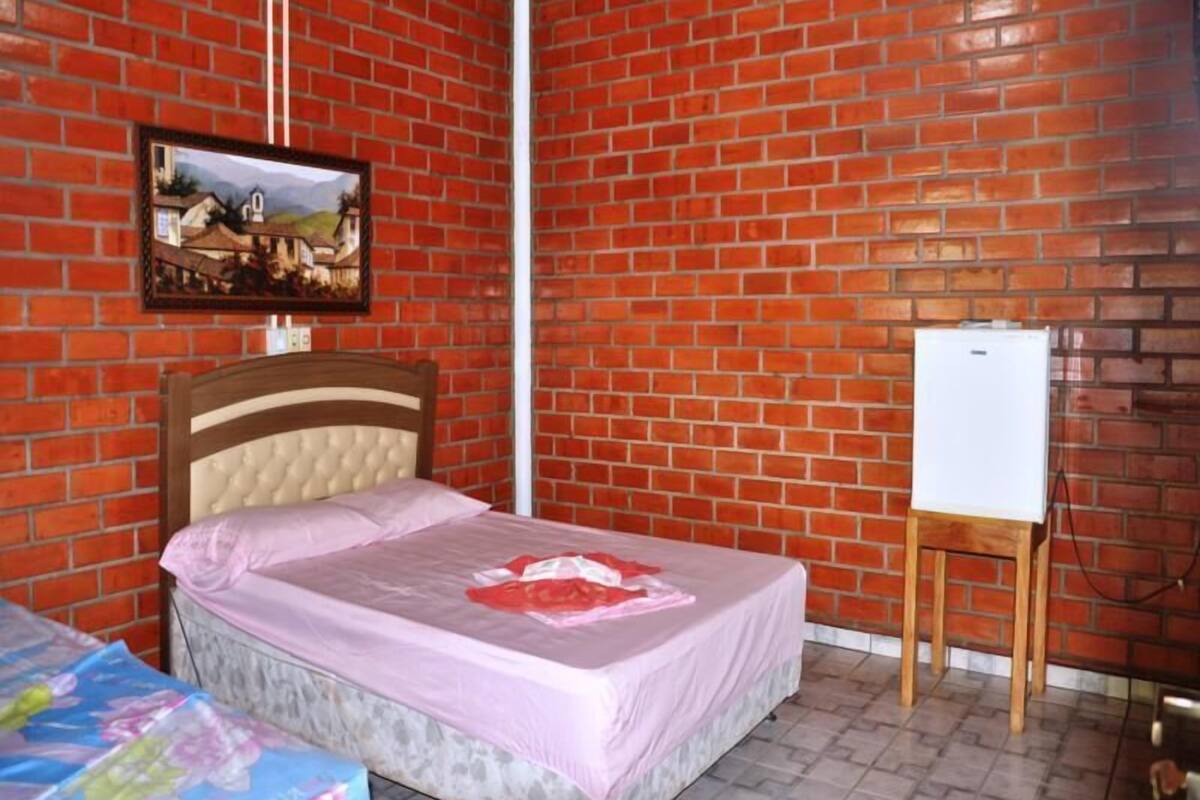 Um quarto com parede de tijolos, uma cama com cabeceira, uma mesa pequena e um quadro branco.