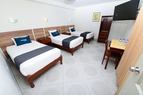 A hotel room with three wooden beds, each with a blue pillow and a white bedspread. There is a wooden desk with a chair and a small table with water bottles.