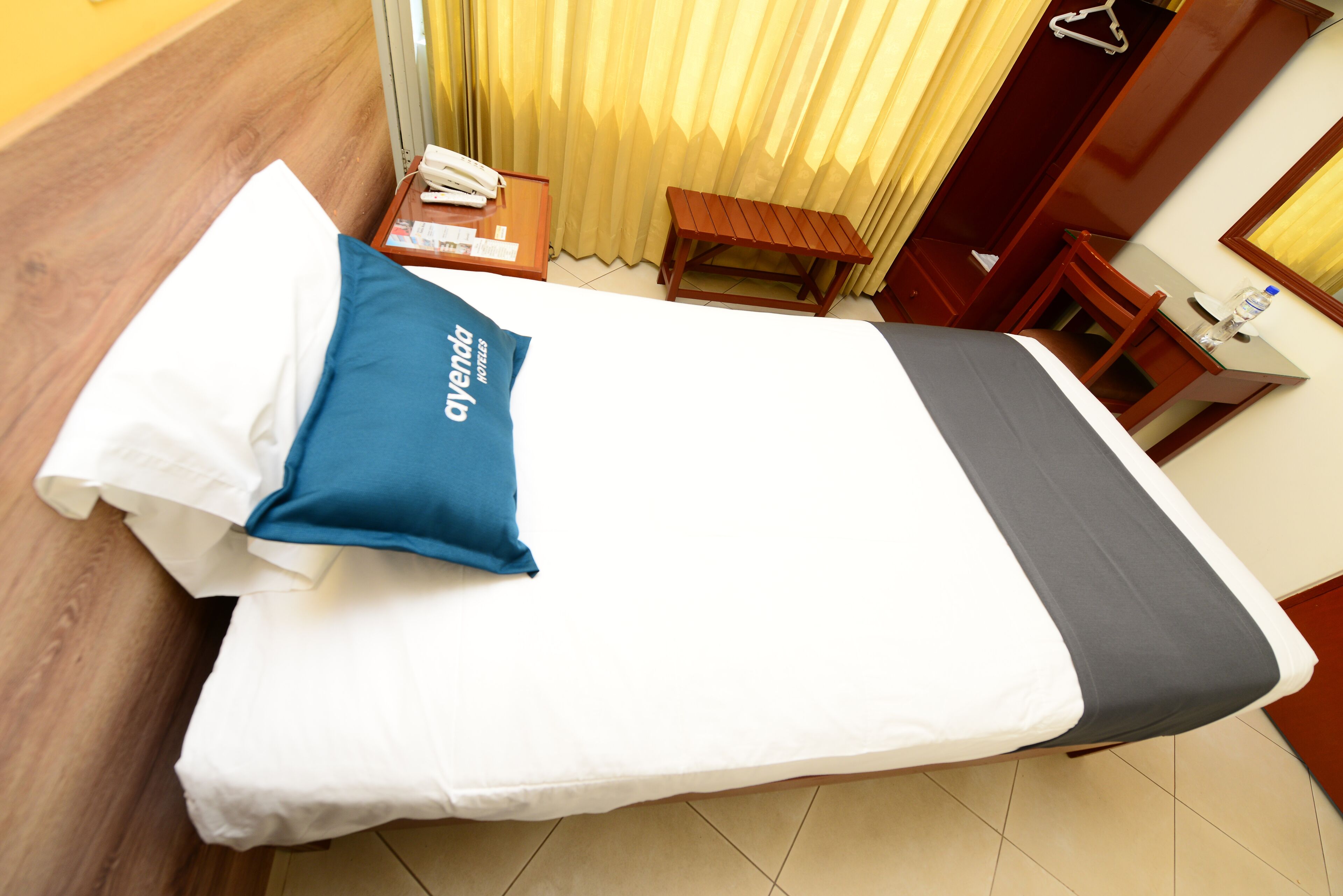 Una cama bien tendida con una colcha blanca y gris, una almohada azul y un cabecero de madera.