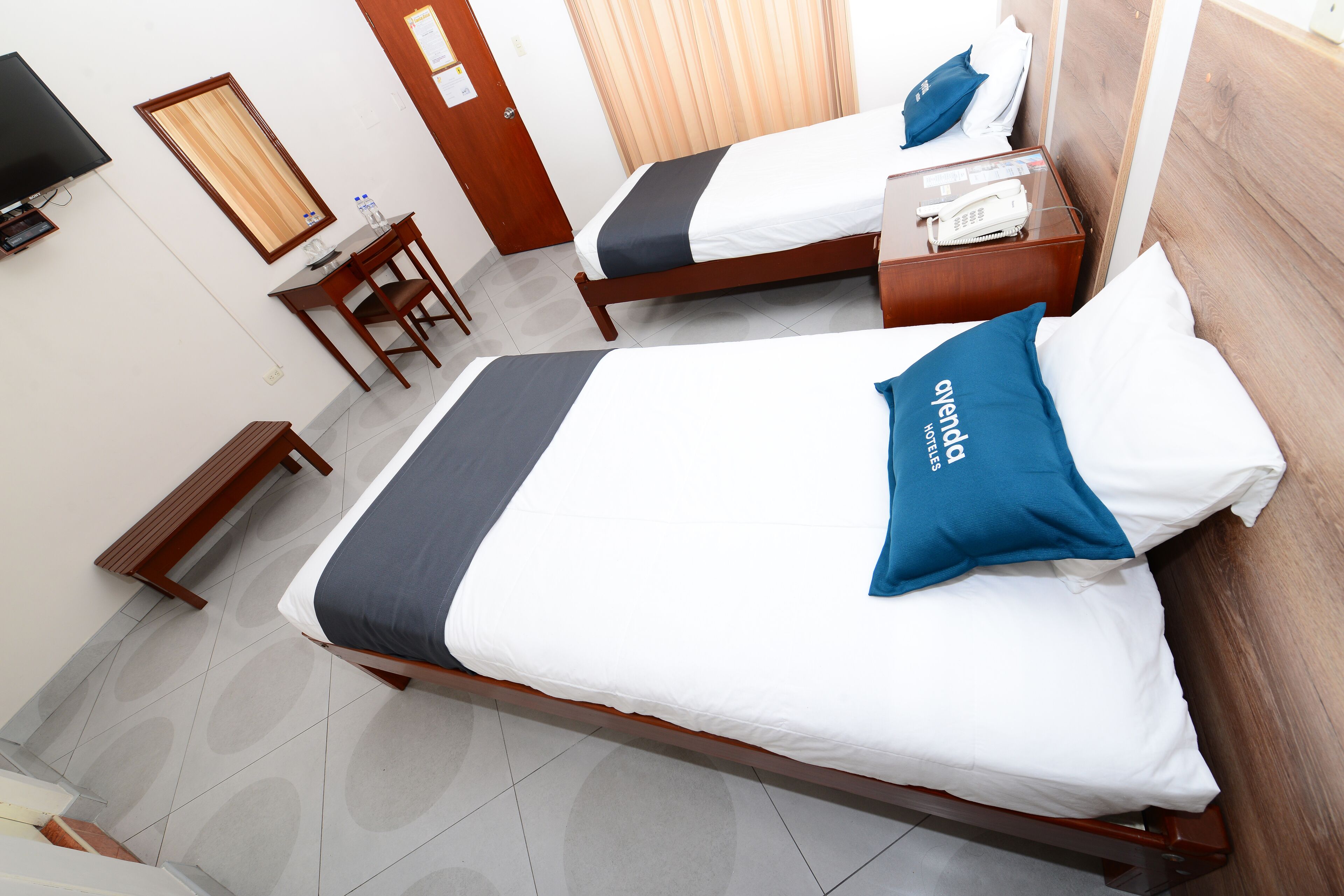 A hotel room with two beds, a desk, a chair, a TV, and a door.