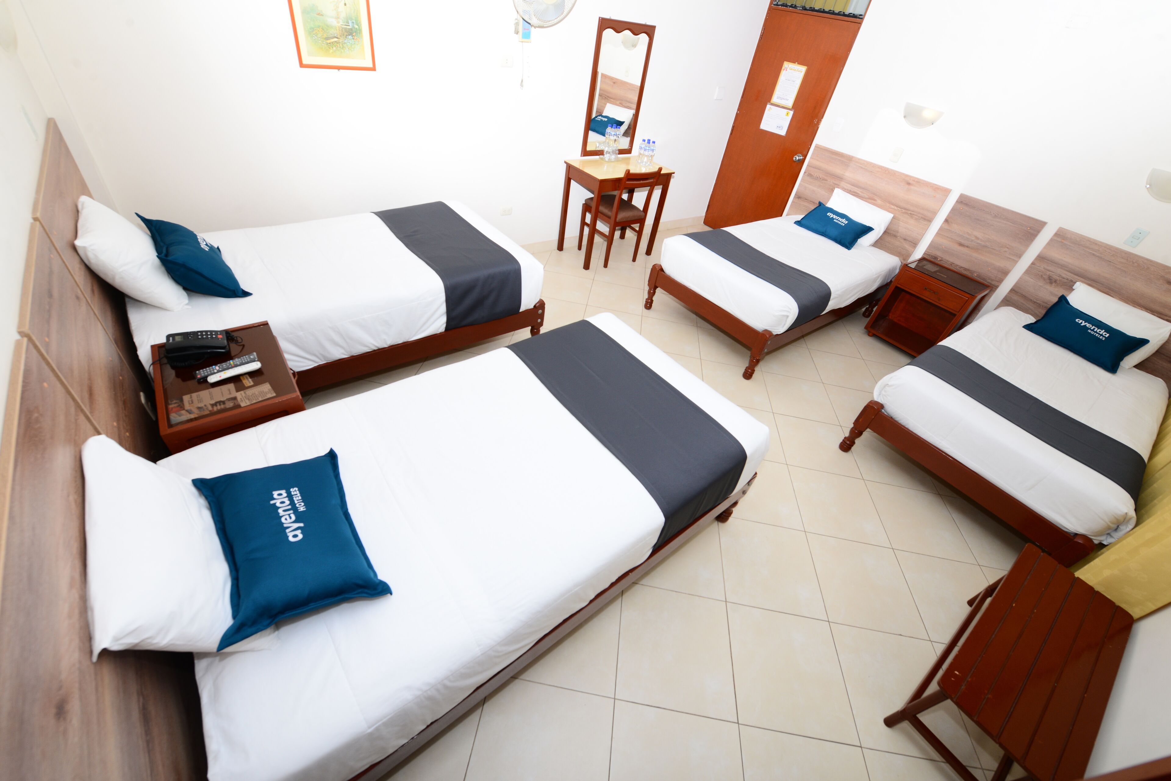 A hotel room with four single beds, each with a blue pillow and a wooden headboard. There is a wooden desk with a chair, a small table with a lamp, and a wooden nightstand with a lamp.