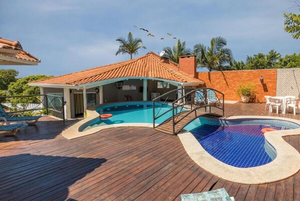 A pool area with a wooden deck, a covered seating area, and a building with a tiled roof.