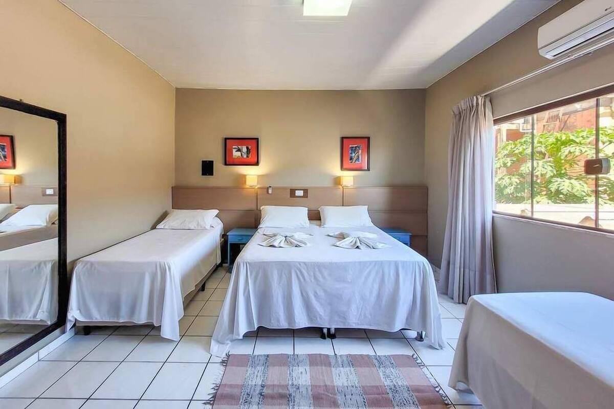 A hotel room with two single beds, a larger bed, a mirror, two bedside tables, and a window with curtains.