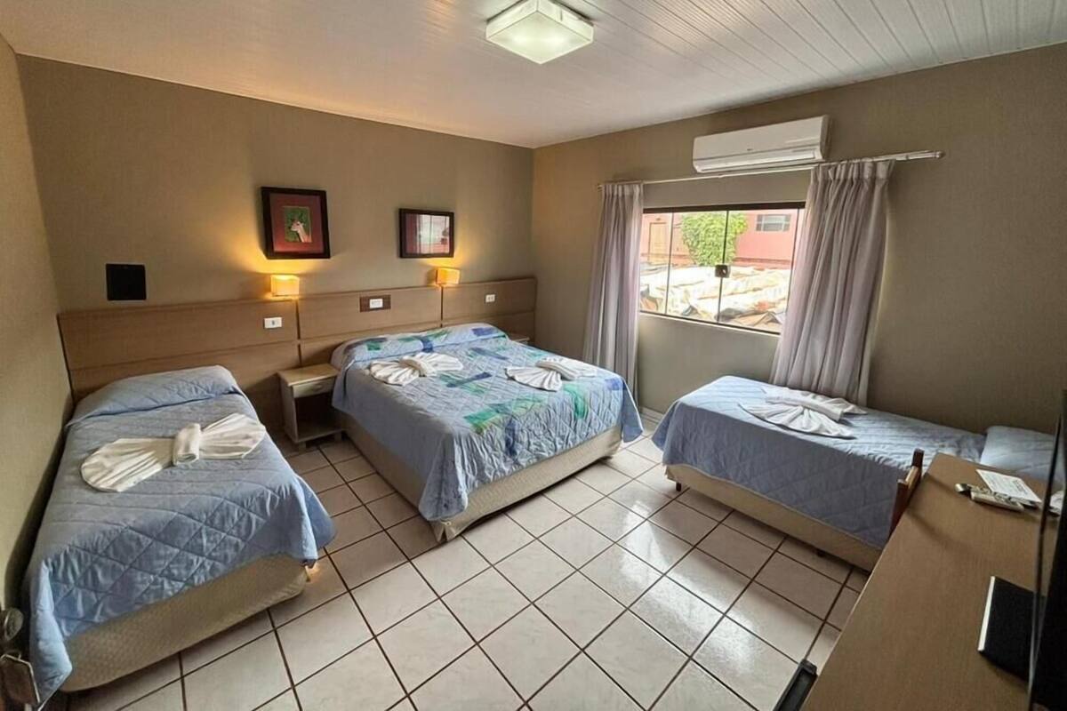 A hotel room with two single beds, a small bedside table with a lamp, a window with curtains, and an air conditioning unit.