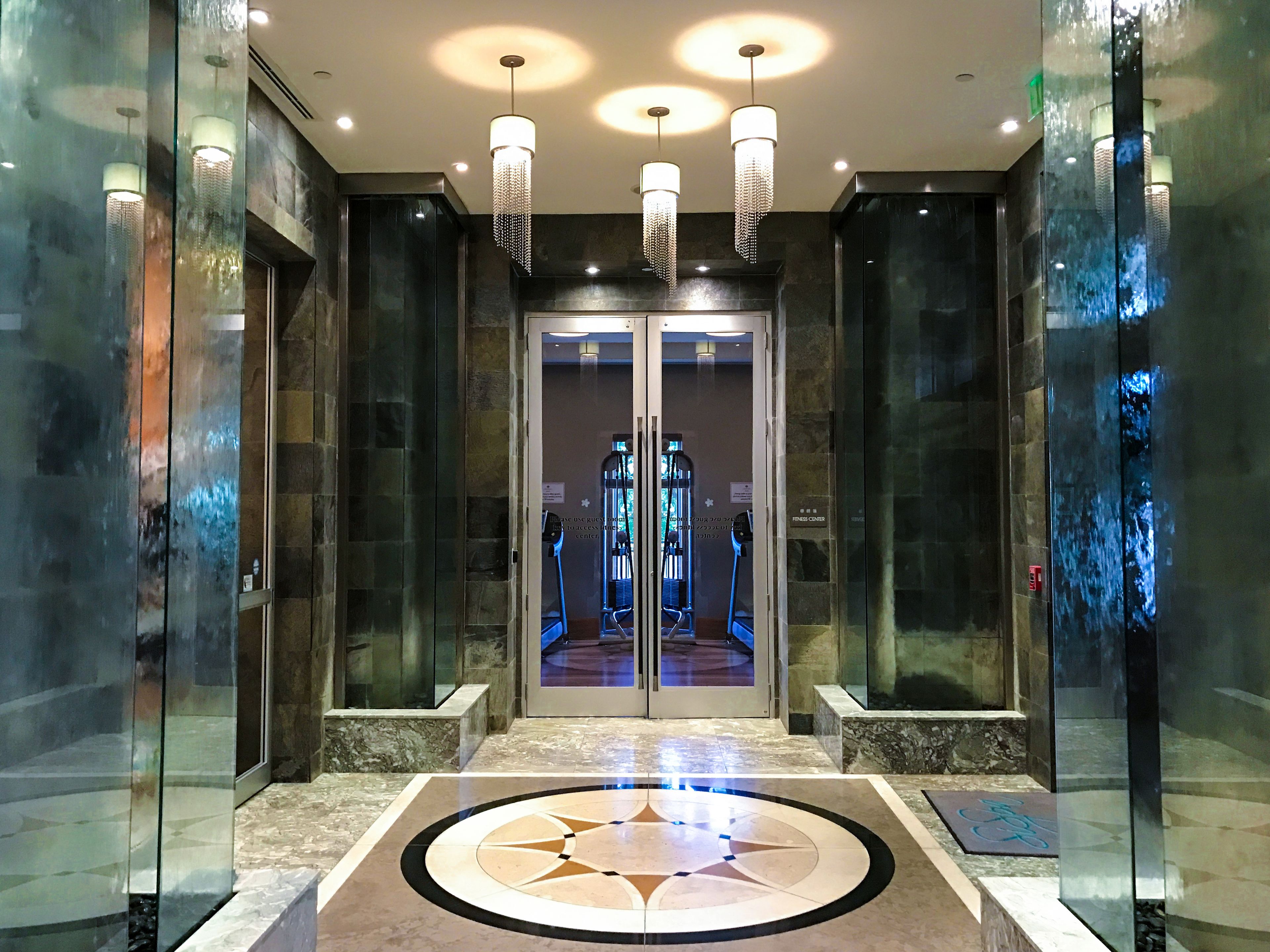 A modern elevator lobby with glass doors, a geometric floor design, and three chandeliers.