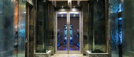 A modern elevator lobby with glass doors, a geometric floor design, and three chandeliers.