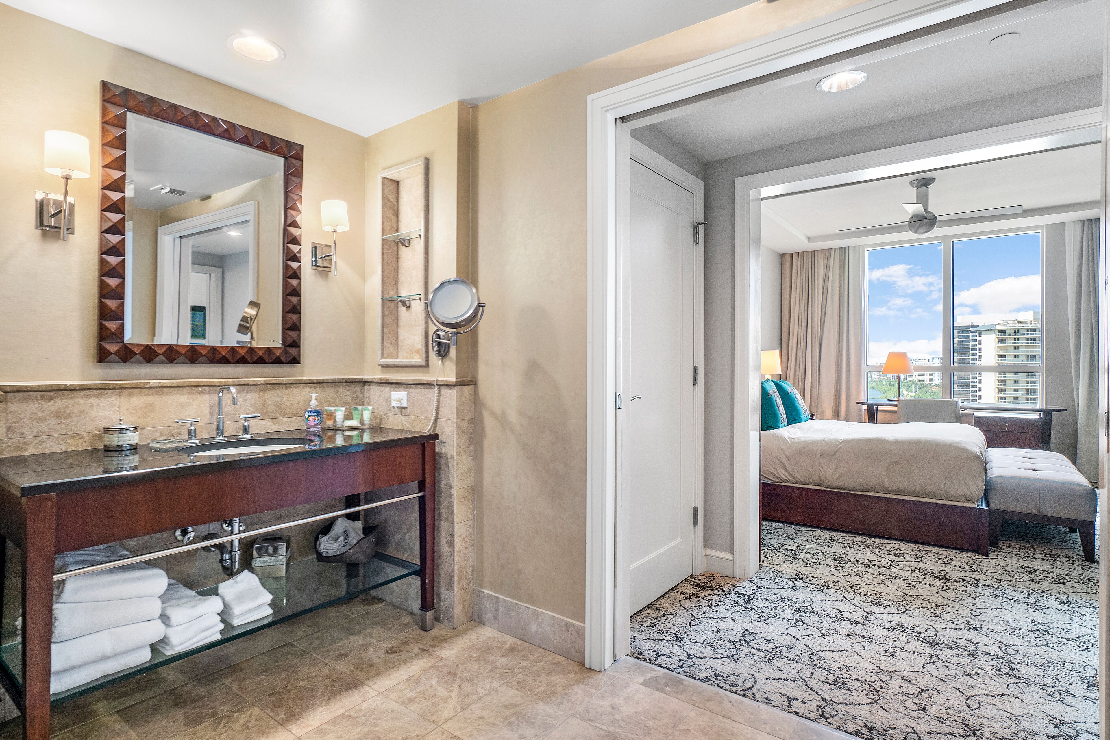 A hotel bathroom with a large mirror, vanity, and a bed in the adjacent room.