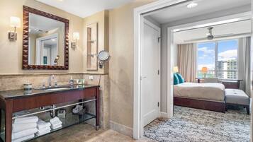 A hotel bathroom with a large mirror, vanity, and a bed in the adjacent room.