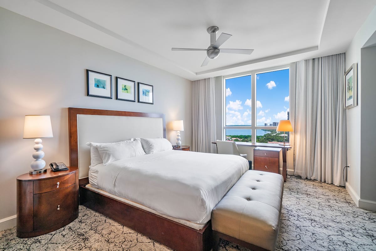 Habitación de hotel con una cama grande, un escritorio con silla, un ventilador de techo y vistas a la ciudad a través de amplias ventanas.