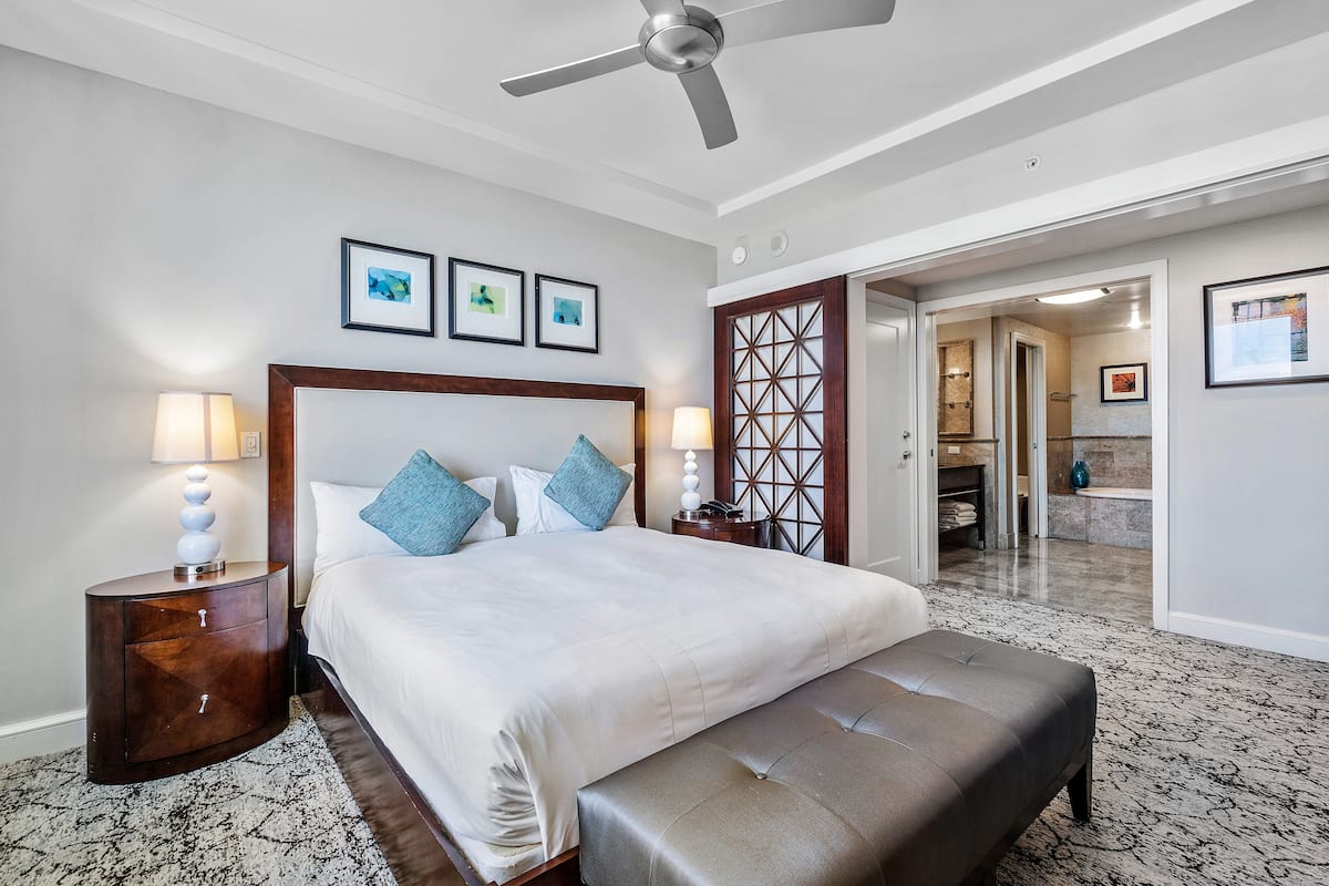Habitación de hotel con una cama grande, un banco, ventilador de techo y un baño visible a través de una puerta abierta.