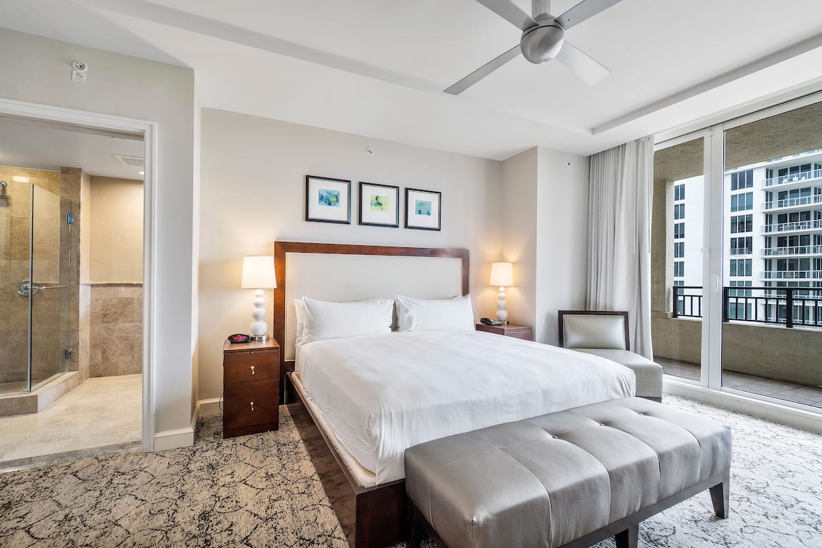 Habitación de hotel con una cama grande, un banco, ventilador de techo, baño con ducha de vidrio y balcón con vista a la ciudad.