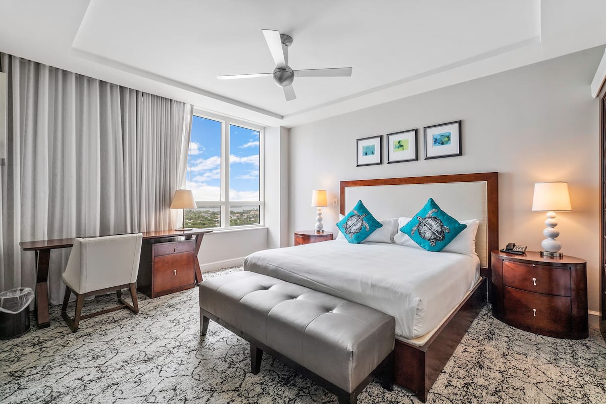 Habitación de hotel con cama, escritorio, silla, banco, ventilador de techo y ventana con cortinas.