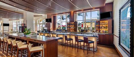 A modern bar area with a long counter, multiple bar stools, and a large window offering a view of palm trees.