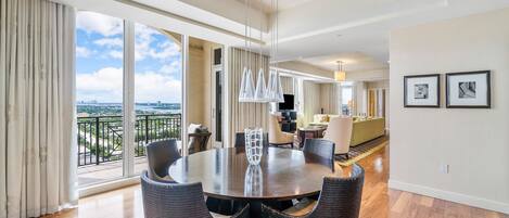 A spacious dining area with a round table and wicker chairs, large windows with a city view, and a modern ceiling design.