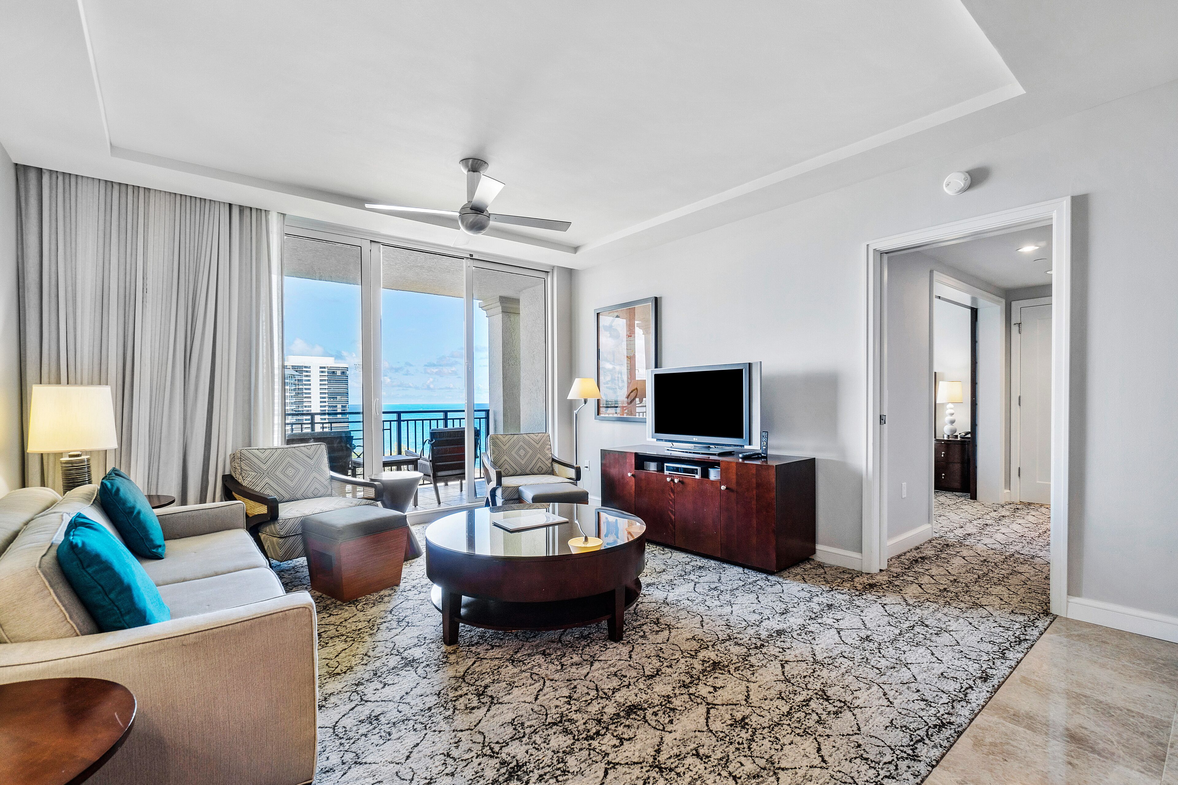 A modern hotel room with a living area featuring a sofa, armchairs, a coffee table, and a flat-screen TV. The room has a large window with a view of the cityscape.