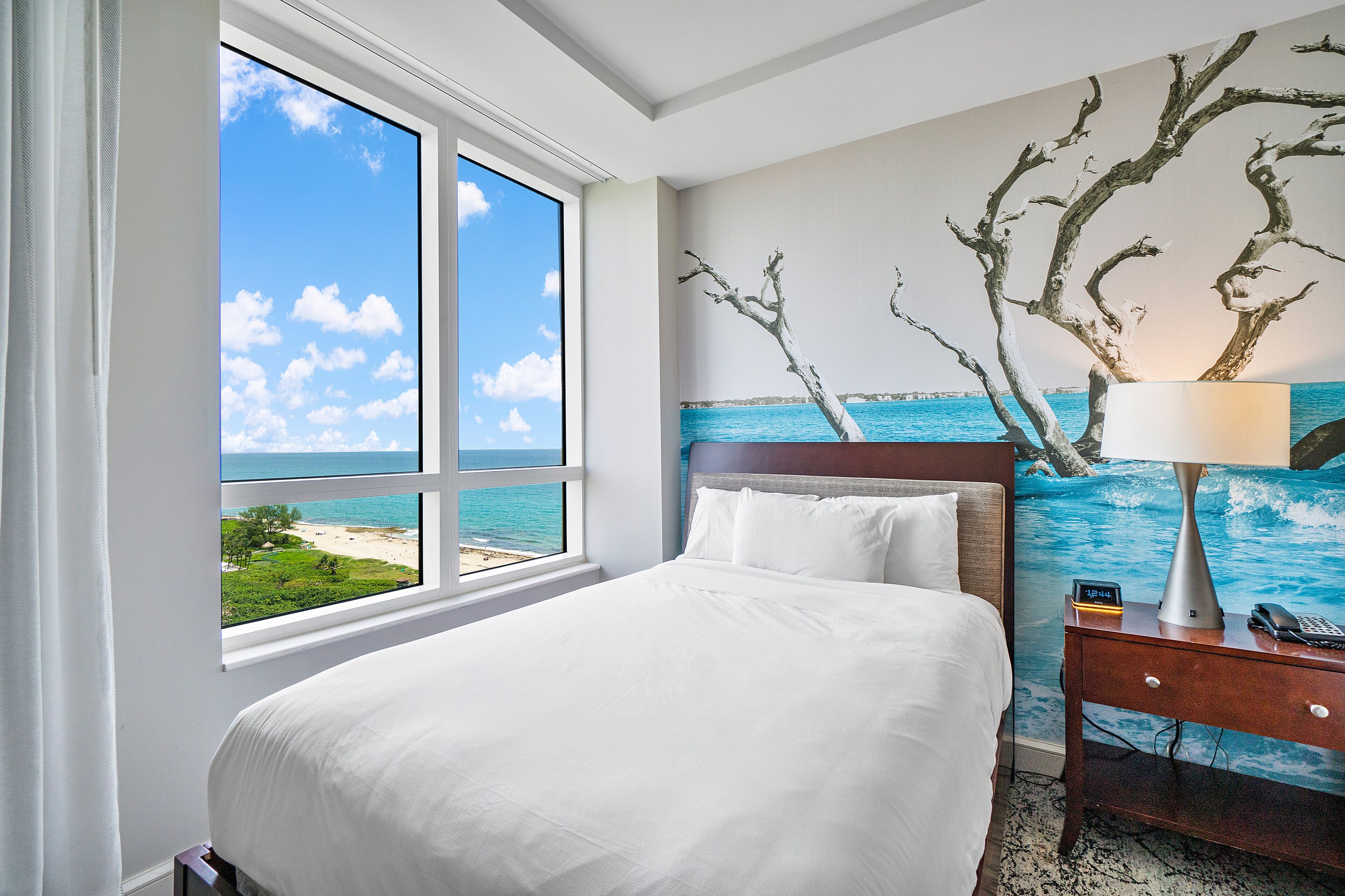 A hotel room with a bed, a nightstand with a lamp, and a view of the beach through a large window.