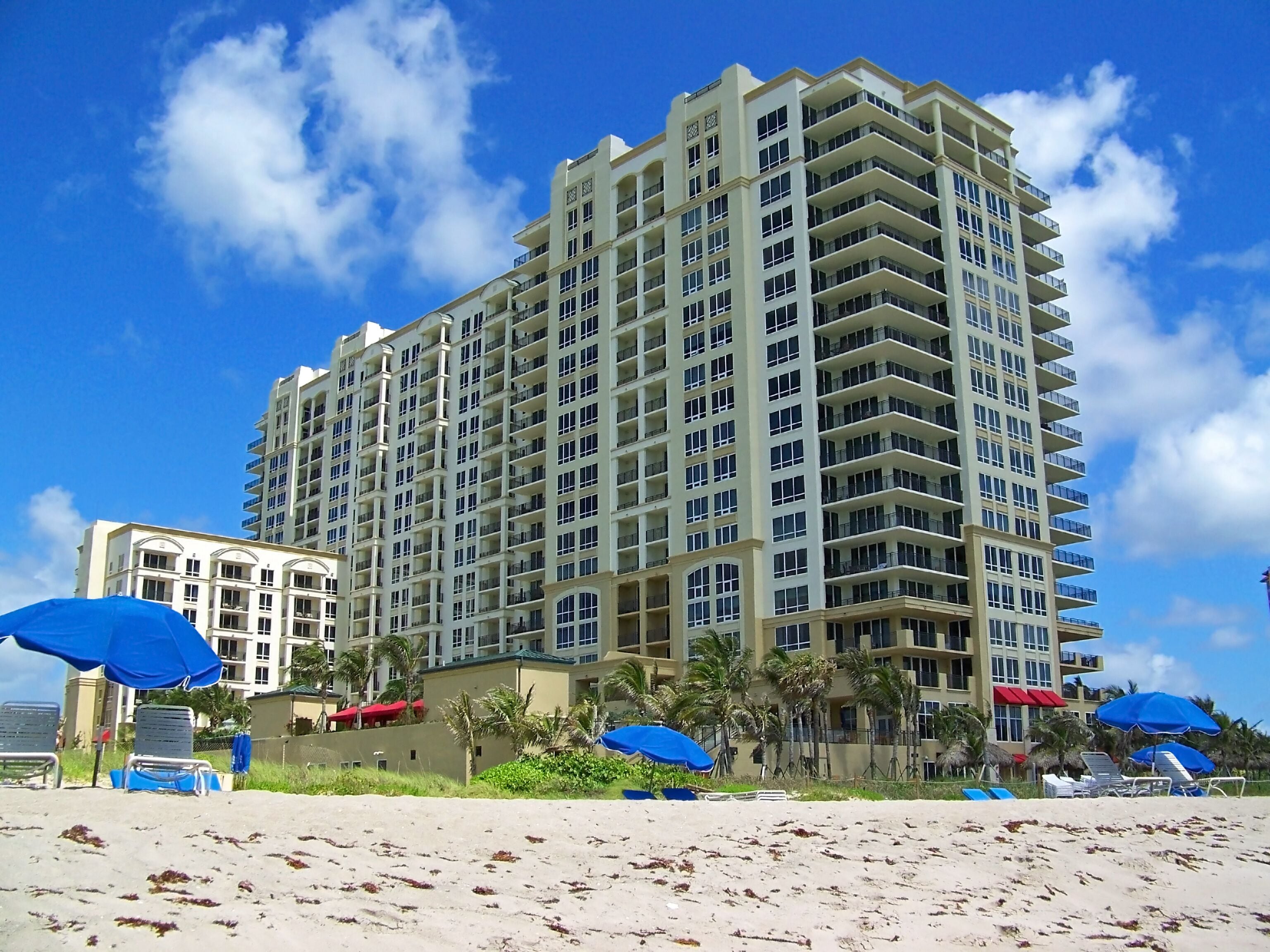 Hotel alto com várias varandas, situado perto de uma praia com guarda-sóis e espreguiçadeiras.