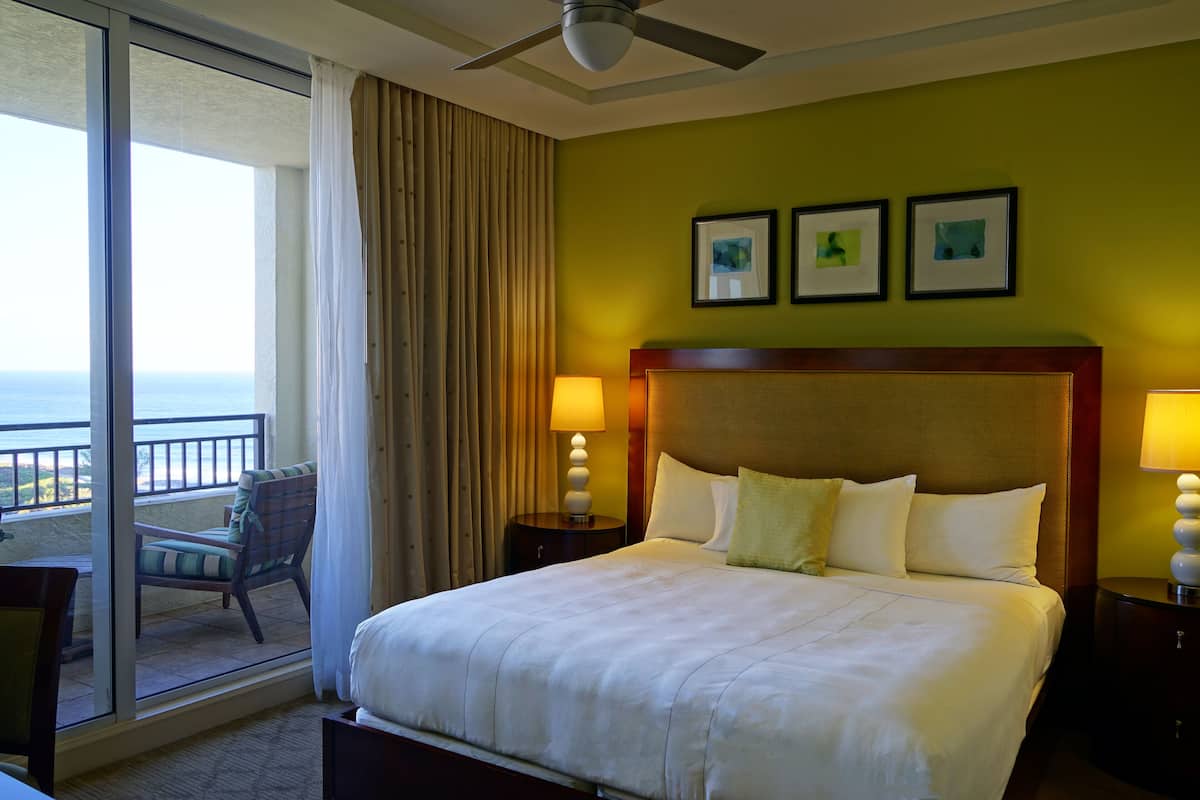 Habitación de hotel con una cama grande, dos lámparas de noche, vista al océano a través de una puerta corrediza y una silla cerca de la puerta.