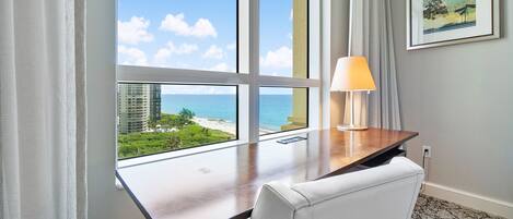 A hotel room with a large window offering a view of the beach and ocean, a wooden desk with a lamp, and a framed picture on the wall.