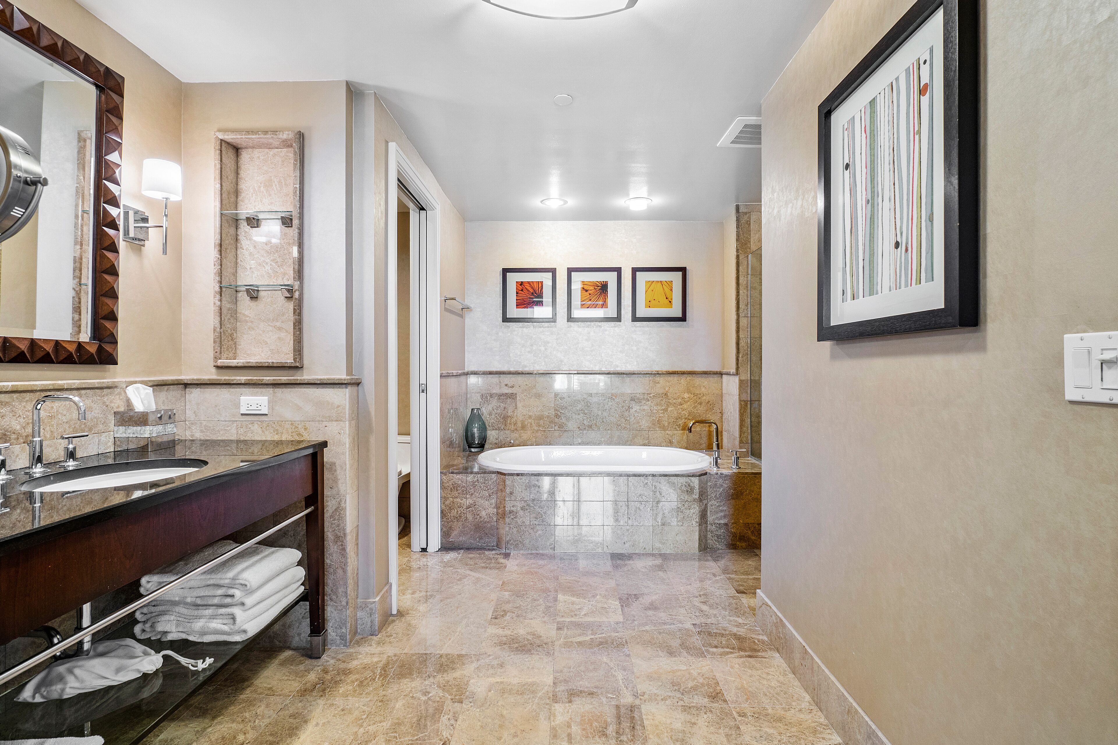 A modern bathroom with a large sink, vanity, and bathtub. There are framed artworks on the wall and a mirror with a geometric frame.