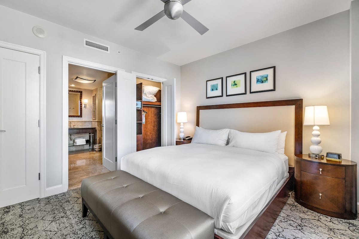 Una habitación de hotel con una cama grande, un banco, una mesita de noche con lámparas y un ventilador de techo.
