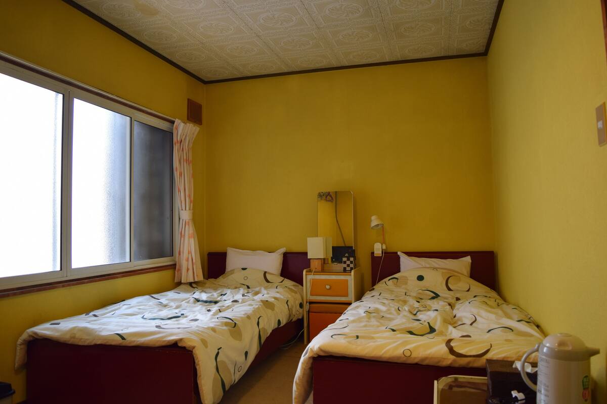 A hotel room with two beds, a window with curtains, a nightstand with a lamp, and a coffee maker.