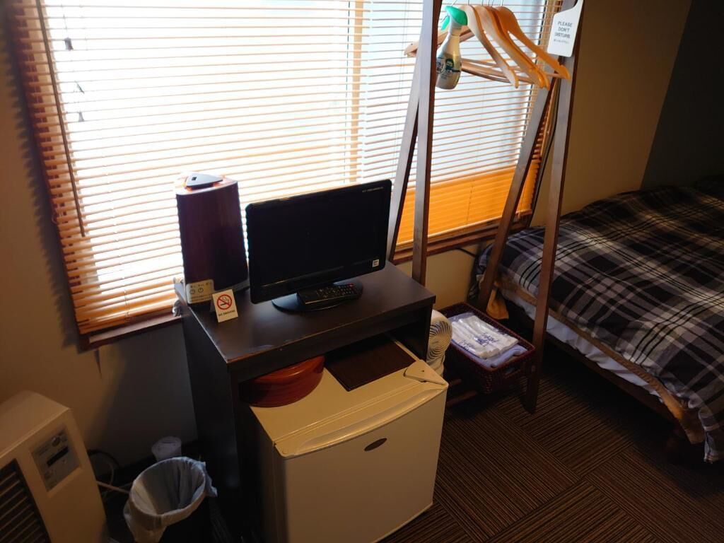 A compact hotel room with a bed, a desk with a computer, a small heater, and a window with blinds.