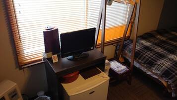 A compact hotel room with a bed, a desk with a computer, a small heater, and a window with blinds.