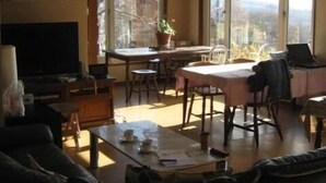 A living room with a sofa, a TV, a dining table with chairs, and large windows offering a view of the outside.