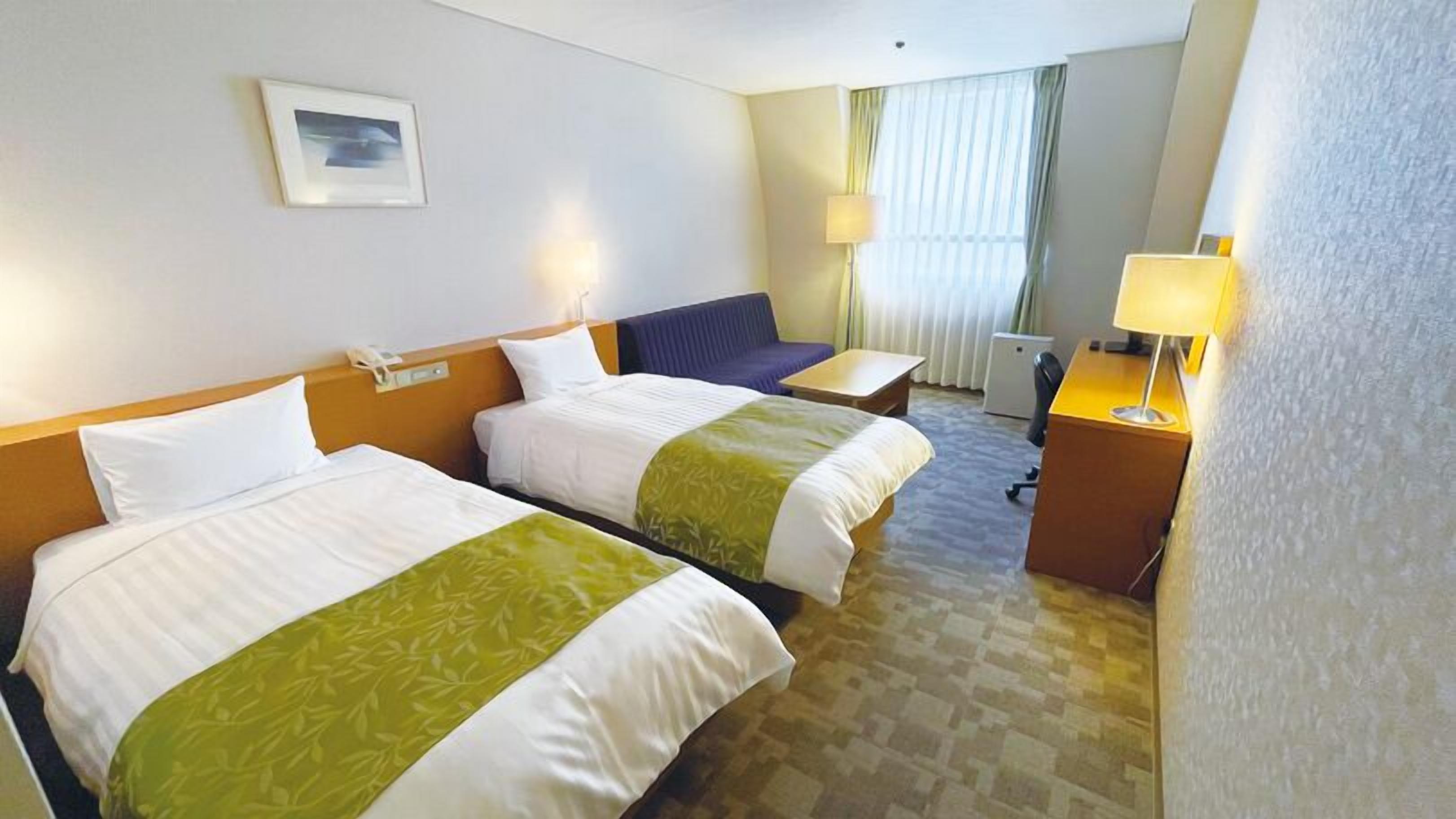 A hotel room with two beds, a desk, a chair, a lamp, and a window with curtains.