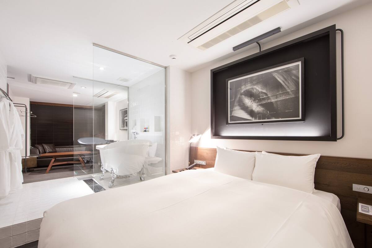 A modern hotel room with a large bed, a glass-enclosed bathroom, a mounted artwork, and a wooden headboard.