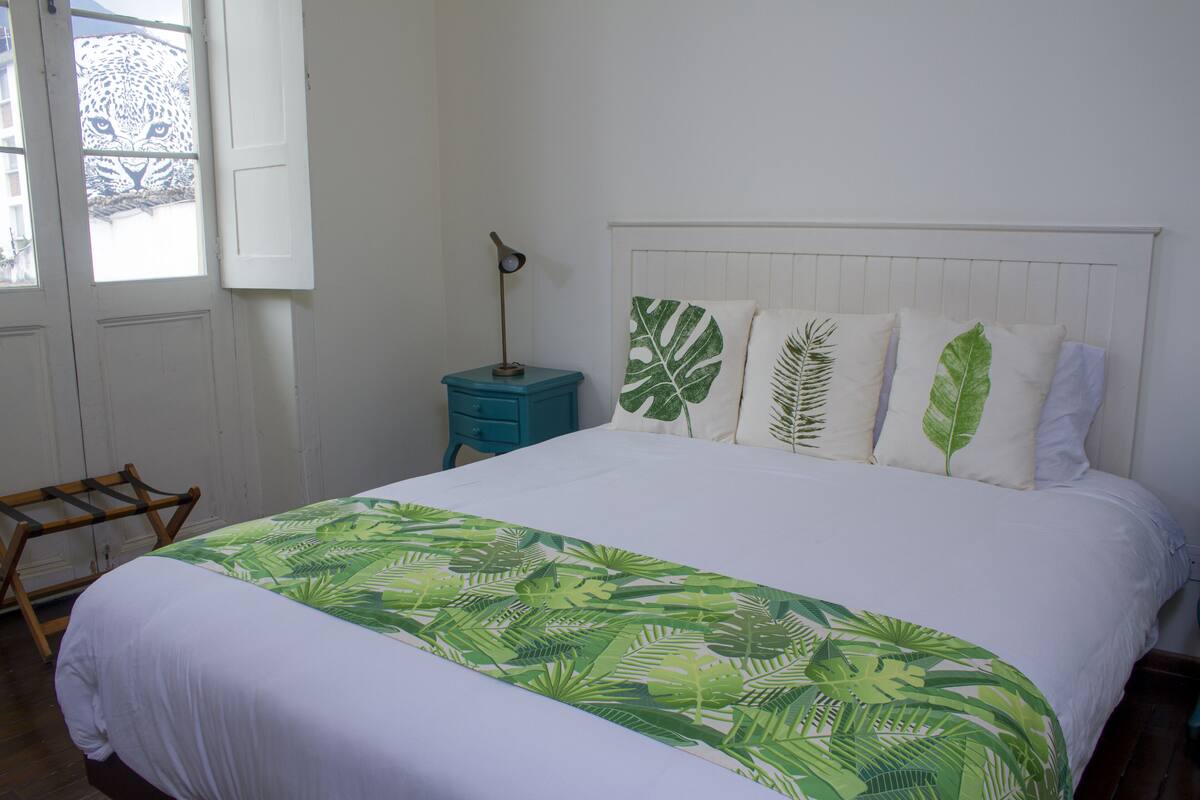 A bed with a white sheet and a green leaf-patterned bedspread. There are three pillows with leaf designs and a nightstand with a lamp.