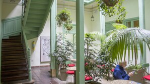 A staircase with green railings, a hanging plant, and a person reading a book in a room with a high ceiling and potted plants. - Hotel Casa De La Vega (Bogotá)