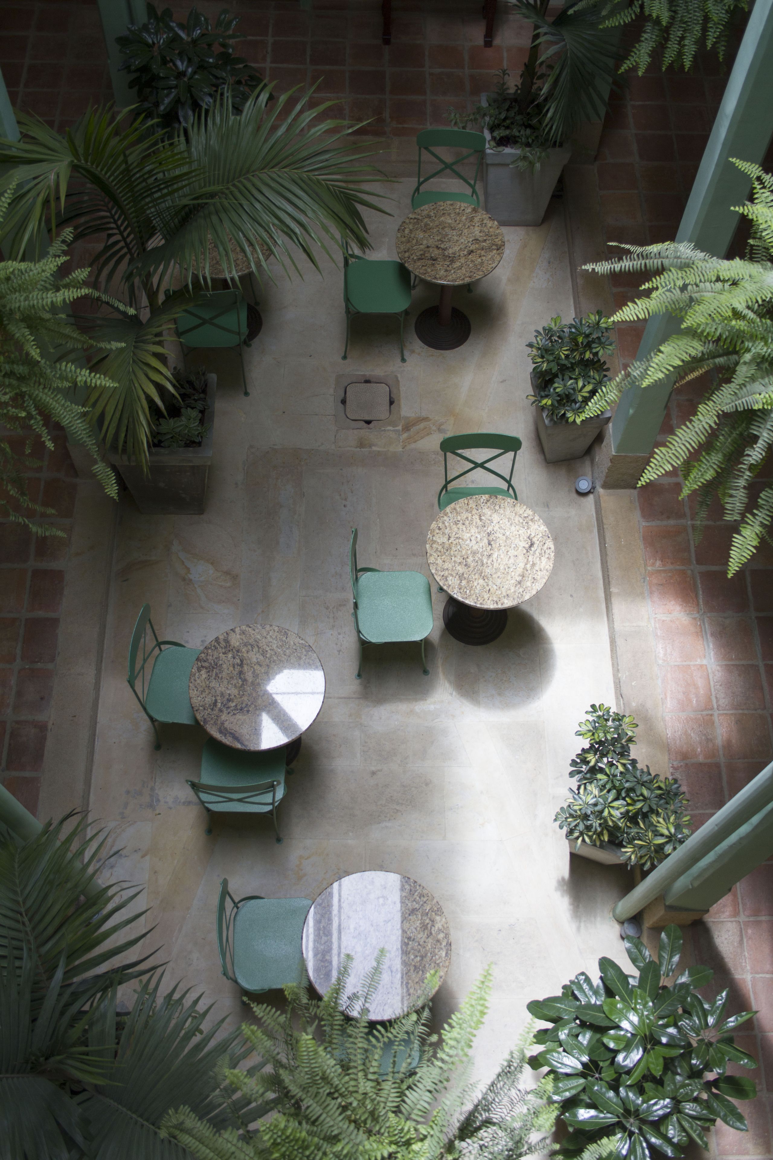 Vue aérienne d’une cour intérieure avec plusieurs tables et chaises rondes, entourée de plantes en pot et de verdure.