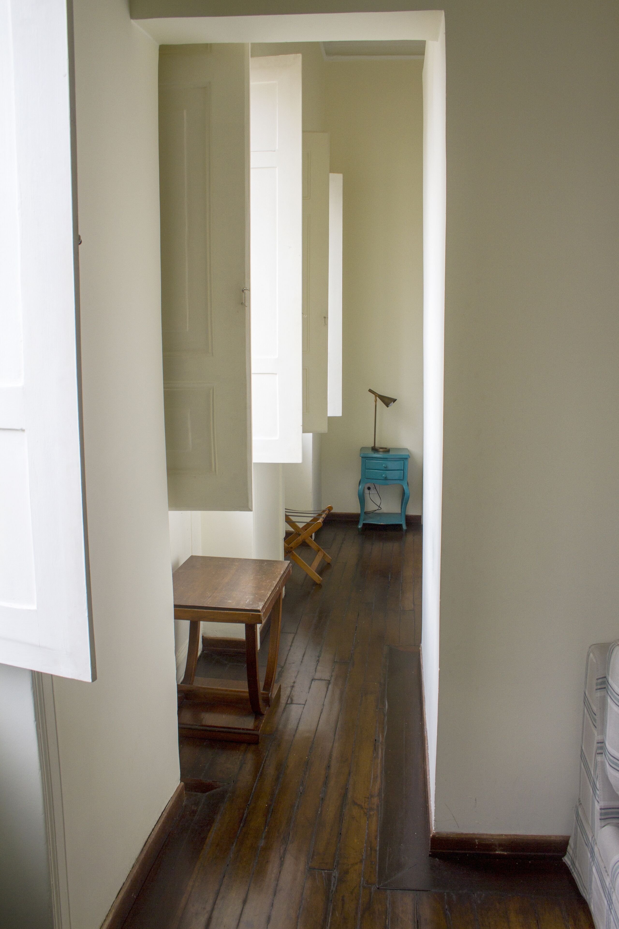 A narrow hallway with wooden flooring, a small wooden table, a blue nightstand, and a lamp.
