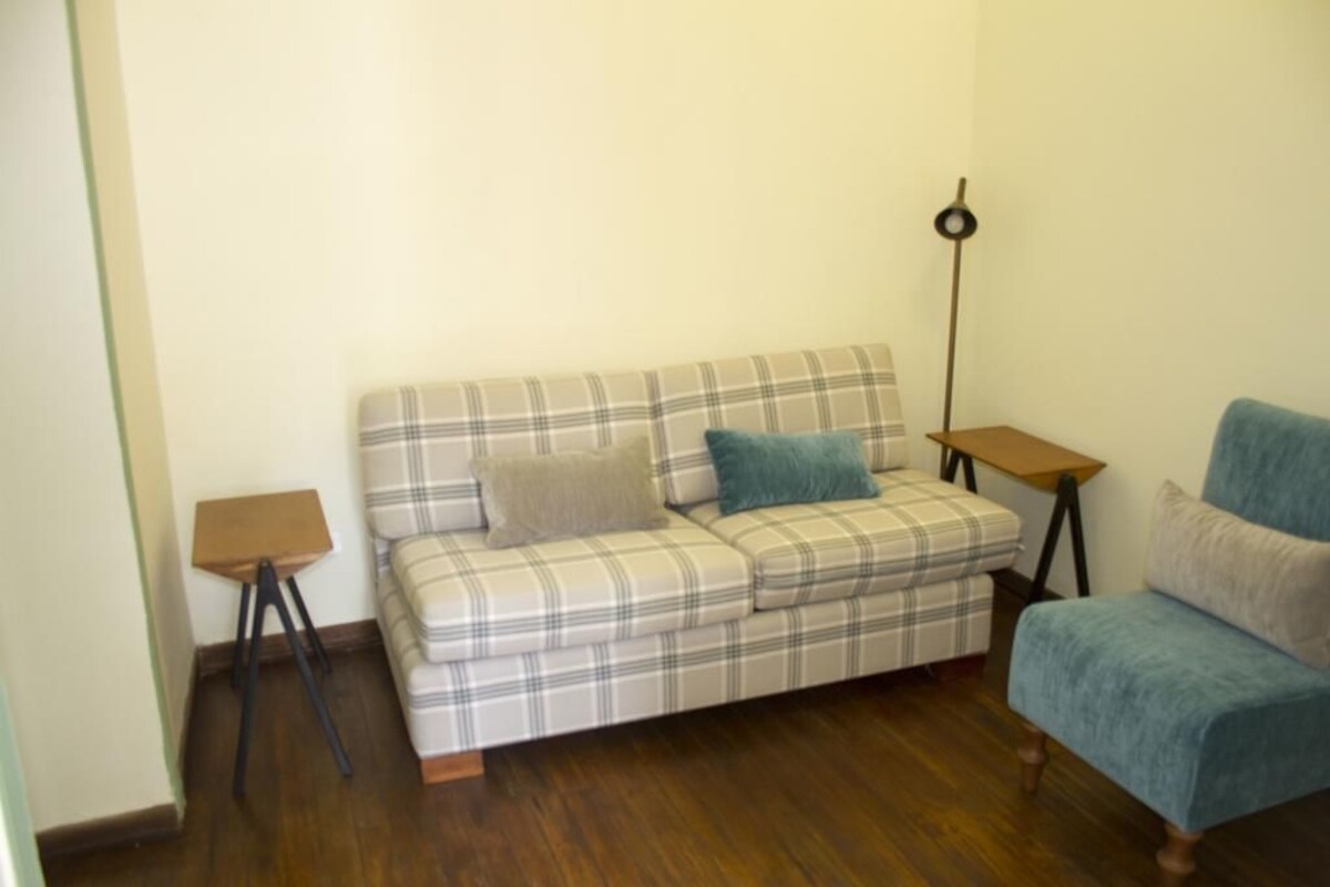 Una habitación con un sofá escocés, un sillón azul, una pequeña mesa de madera y una lámpara de pie.