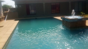 En swimmingpool med en springvand, omgivet af en flisbelagt terrasse og en bygning med et flis tag.