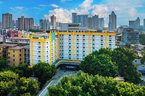 A multi-story hotel with a yellow and white facade, surrounded by greenery and a city skyline in the background. - LNCommunity (Guangzhou)