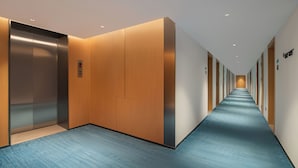 Un pasillo moderno de hotel con alfombra azul, puertas de ascensor de madera y metal, y un largo pasillo con puertas a ambos lados.