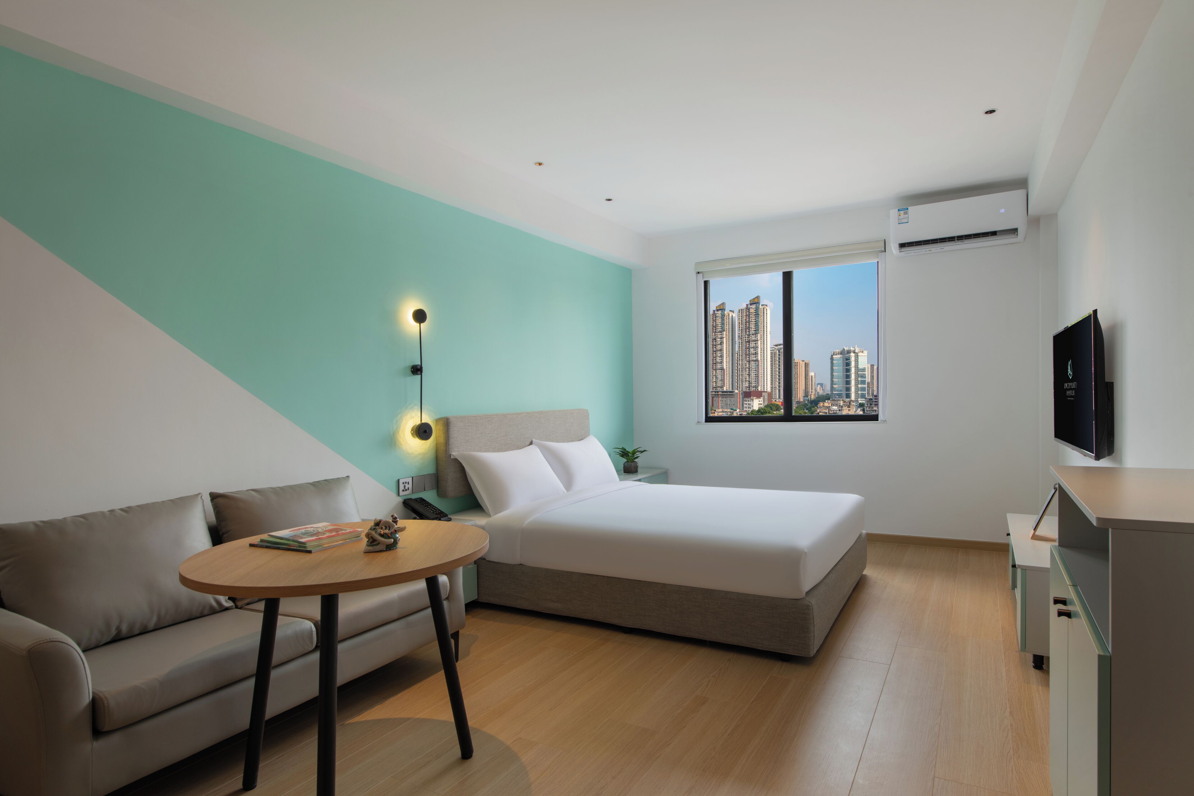 Ein modernes Hotelzimmer mit Bett, einer Sitzecke, einem kleinen Tisch, einem Fernseher und einem Fenster mit Blick auf die Stadt.
