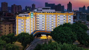 Un hotel de varios pisos con una fachada llamativa en amarillo y blanco, rodeado de abundante vegetación y un horizonte urbano de fondo.