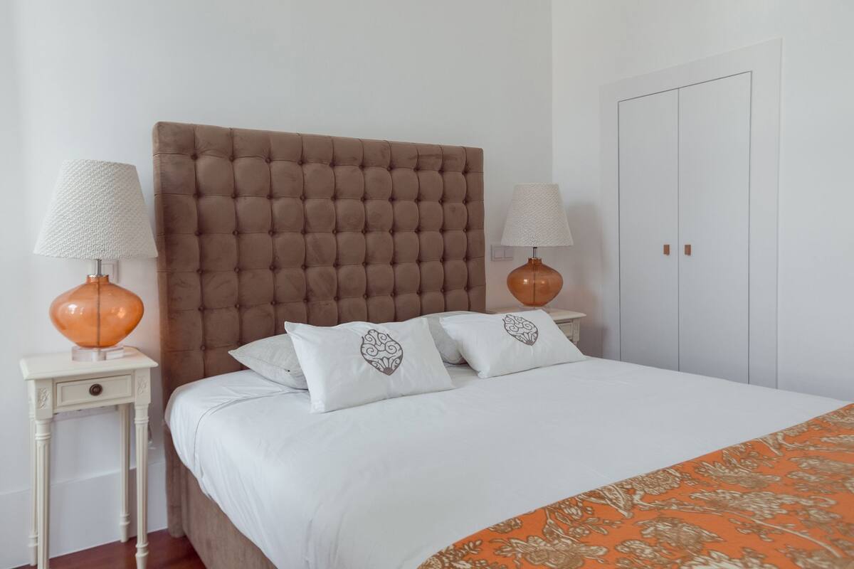 Une chambre à coucher avec une tête de lit capitonnée, du linge de lit blanc, deux tables de chevet avec des lampes et une armoire.