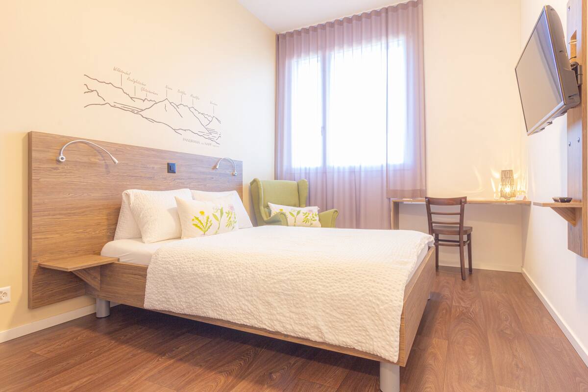 Quarto com cabeceira de madeira, cama com roupa de cama branca, uma cadeira verde, uma escrivaninha com cadeira e uma TV fixada na parede.