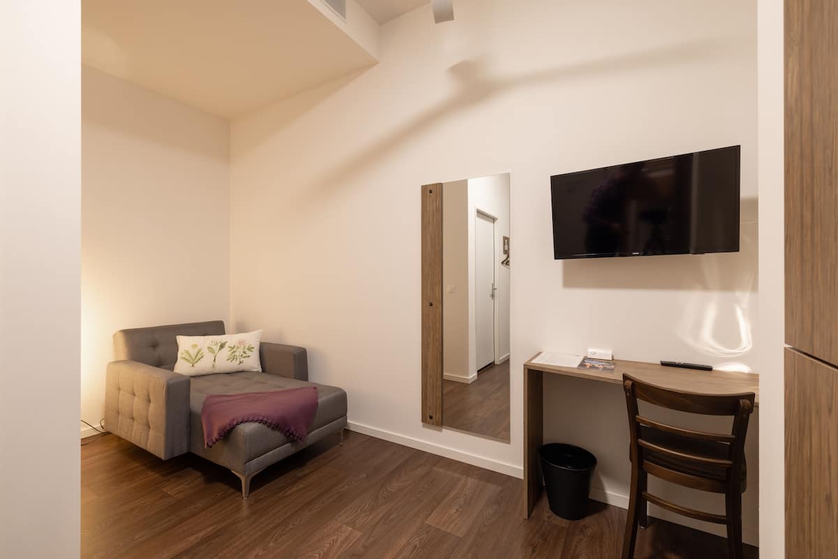Quarto pequeno e moderno com um sofá cinzento, uma secretária de madeira, uma cadeira, uma TV na parede e um espelho.